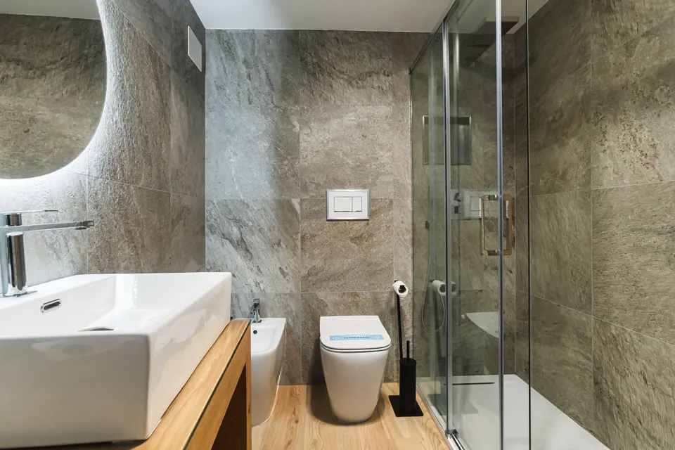 Il bagno della Luna: le pareti di pietra chiara, la doccia di vetro, lo specchio rotondo illuminato e il lavabo su un piano di legno.