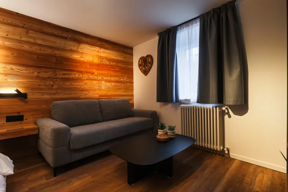 L'angolo del divano letto nel Sole: il divano grigio, un tavolino nero, la parete di larice illuminata e un cuore di legno appeso.