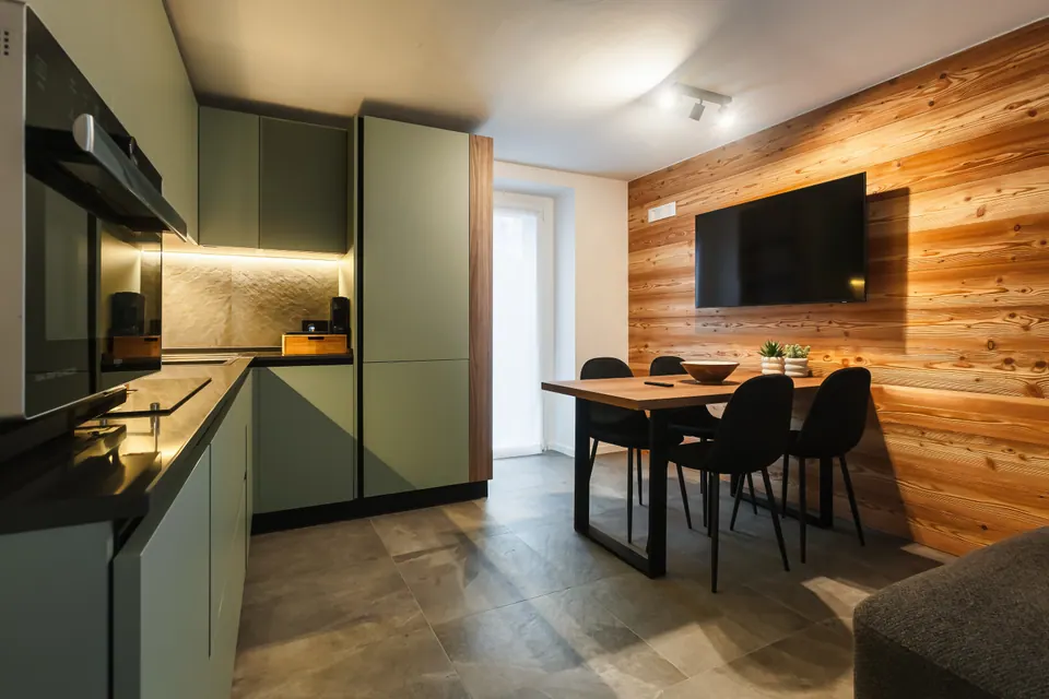 La cucina del Sole con i mobili verde salvia, il tavolo da pranzo con le sedie nere e il televisore sulla parete di larice.