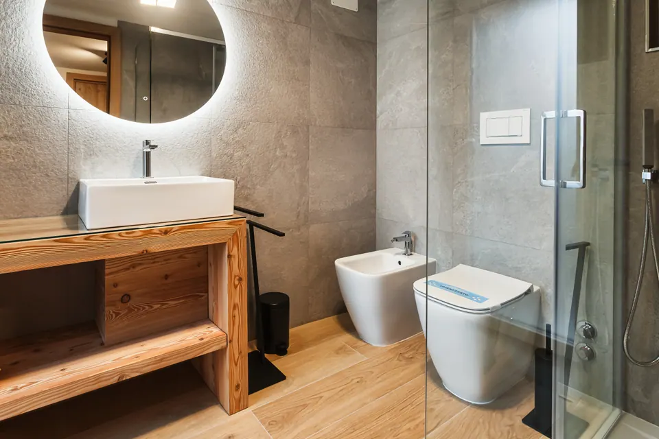Il bagno del Sole: il lavabo su un mobile di legno, lo specchio rotondo illuminato, i sanitari sospesi e la doccia con la parete di vetro.