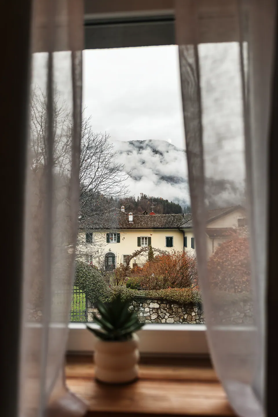 La vista dalla finestra del Sole: le case del paese e le montagne con le nuvole basse, fra le tende leggere.
