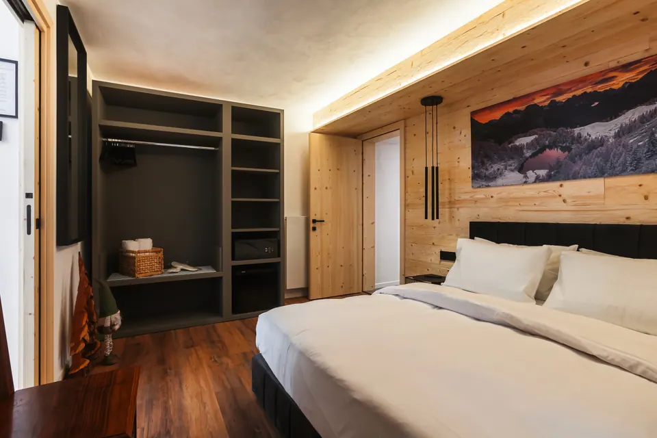 The Tramonto bedroom: a double bed against a spruce wall, a photo of mountains at sunset above the headboard and three pendant lamps; on the left, the open wardrobe.