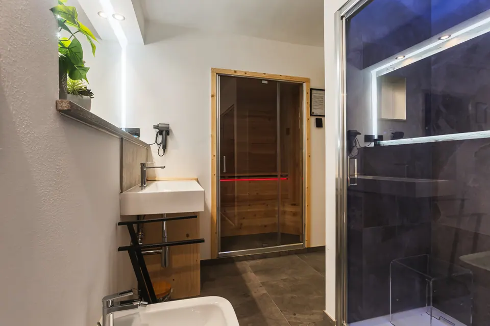 Il bagno del Tramonto con la spa: il lavabo su un mobile di legno, in fondo la sauna con la porta di vetro e a destra il bagno turco con le pareti di pietra scura.