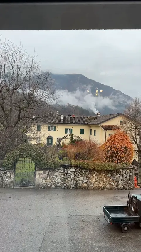 La vista dalla finestra del Sole: una casa gialla, un muro di sassi, gli alberi d'autunno e la montagna con le nuvole basse.